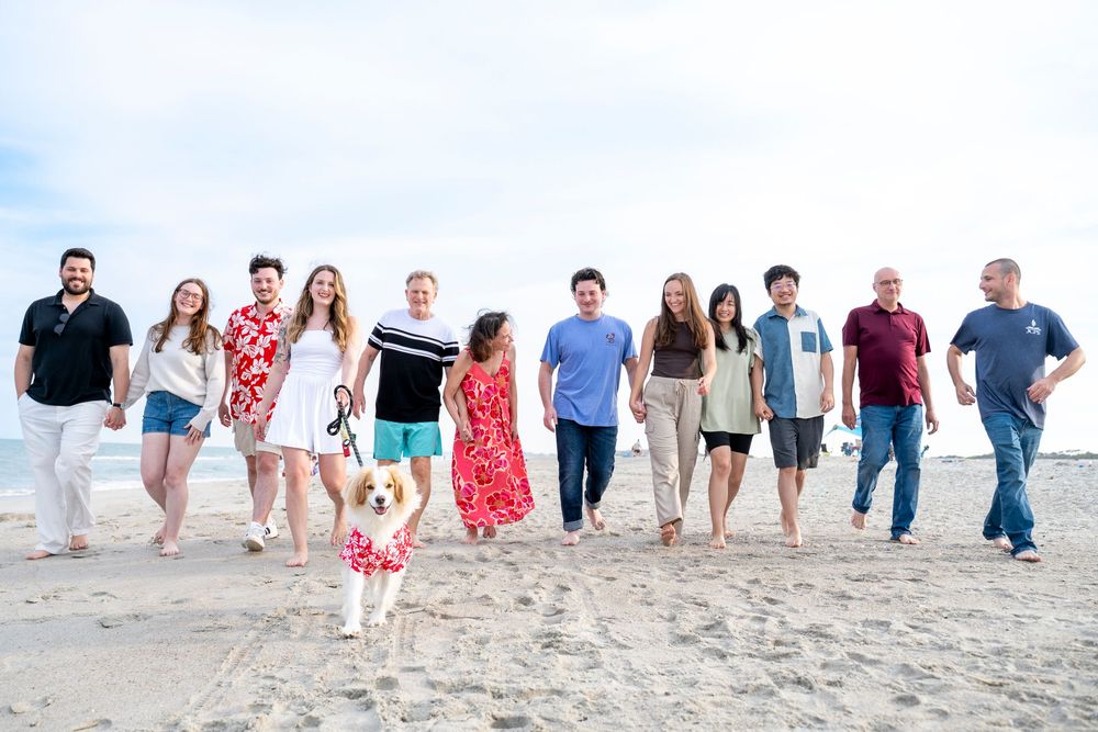 A large group of smiling people and a dog walk together on a sandy beach on a cloudy day.
