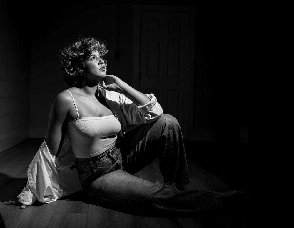 Black and white photo of a woman in a crop top and trousers sitting on a floor, gazing upward dramatically.