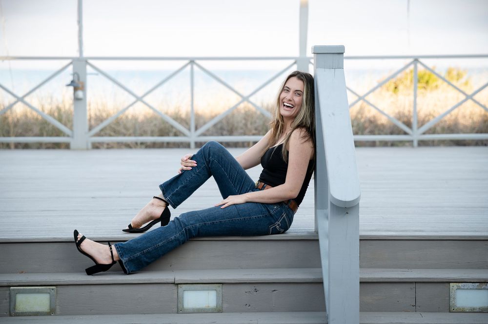 Woman in casual attire sitting relaxed on wooden deck railing with bridge and water in background.