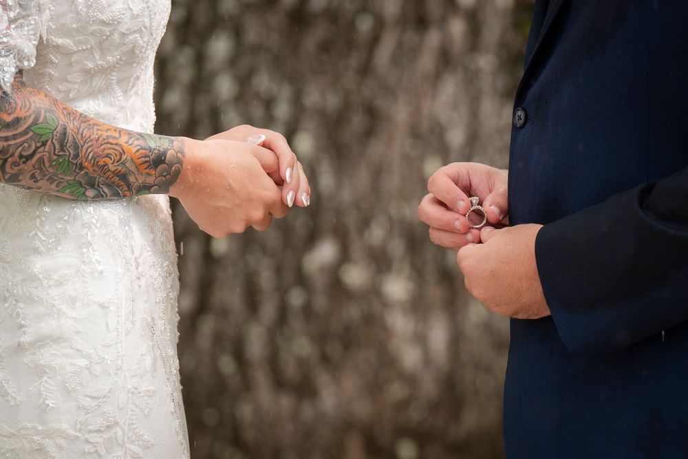 Close up of hands in wedding attire exchanging rings during an outdoor ceremony.