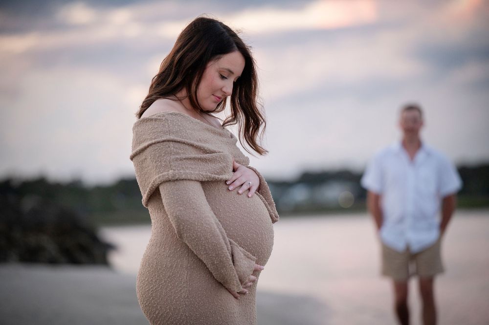 A pregnant woman in a beige sweater dress stands on a beach at sunset while a figure waits in the background.