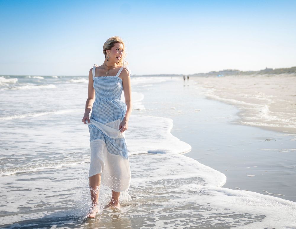 A person in a white dress walks along a sunny beach shoreline as waves gently wash onto the sand.