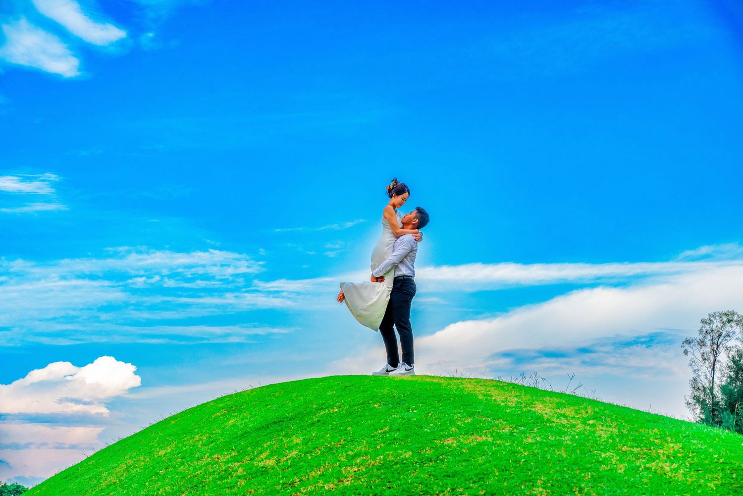 A couple embraces on a grassy hilltop against a bright blue sky.