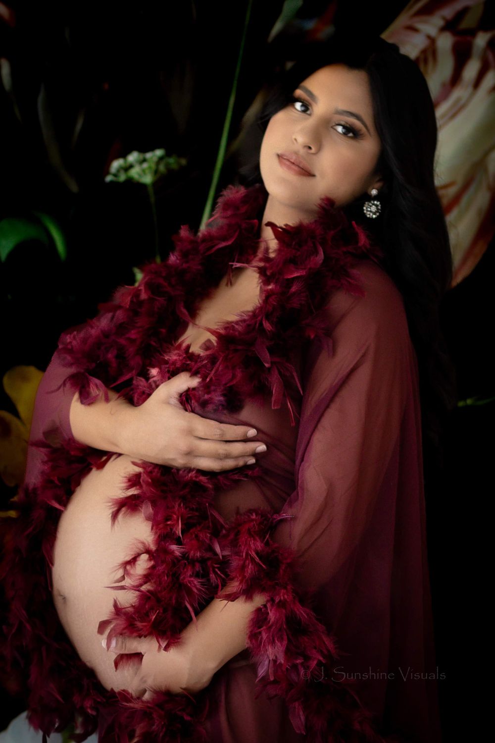 Maternity photo with burgundy feather boa and sheer dark red dress against moody botanical backdrop.