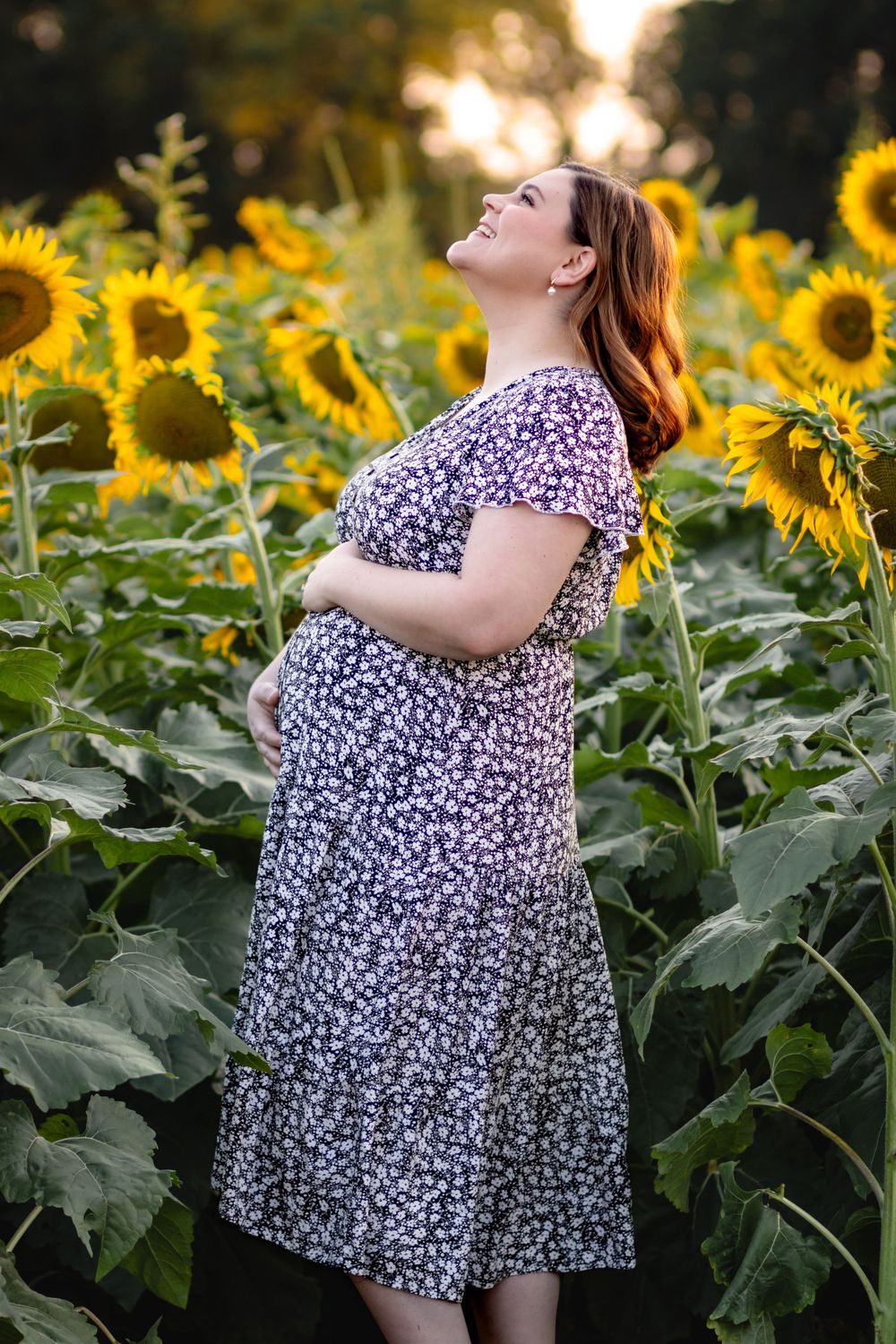 Expecting mother in floral dress poses joyfully in sunflower field during golden hour sunset.