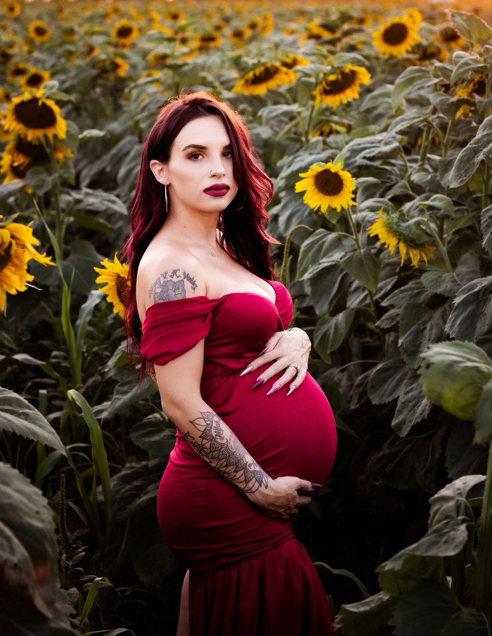 A person in a burgundy off-shoulder dress poses in a sunflower field during golden hour.