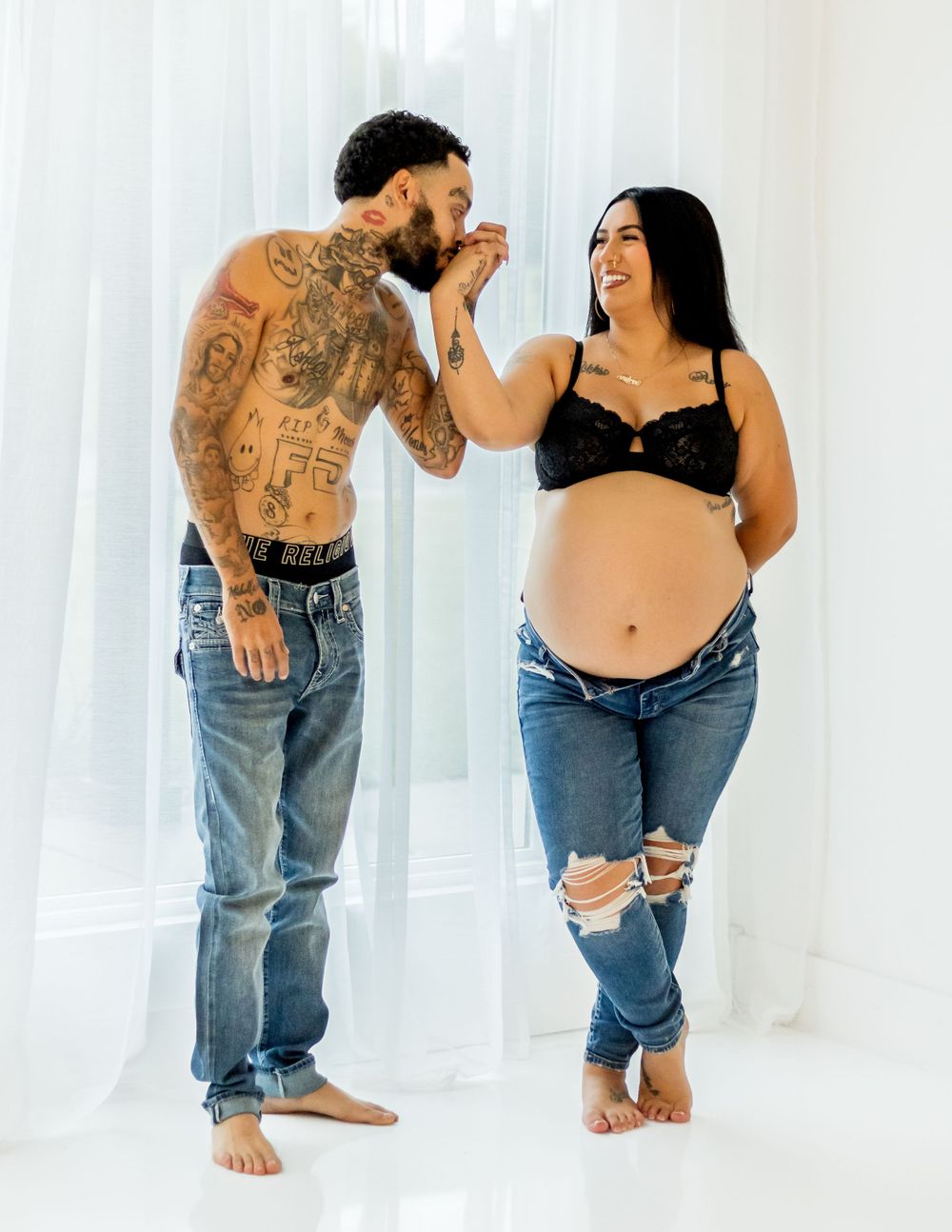 A couple in casual jeans share a sweet moment against a white backdrop during a pregnancy photoshoot.