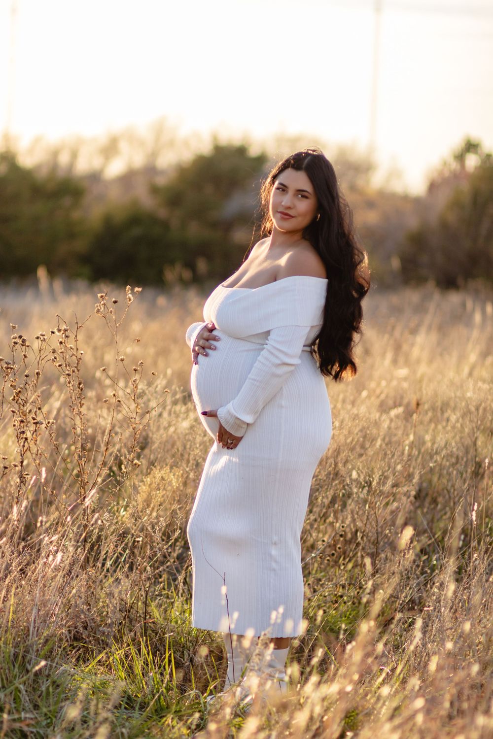 Pregnant woman wearing white off-shoulder dress poses in golden field at sunset.