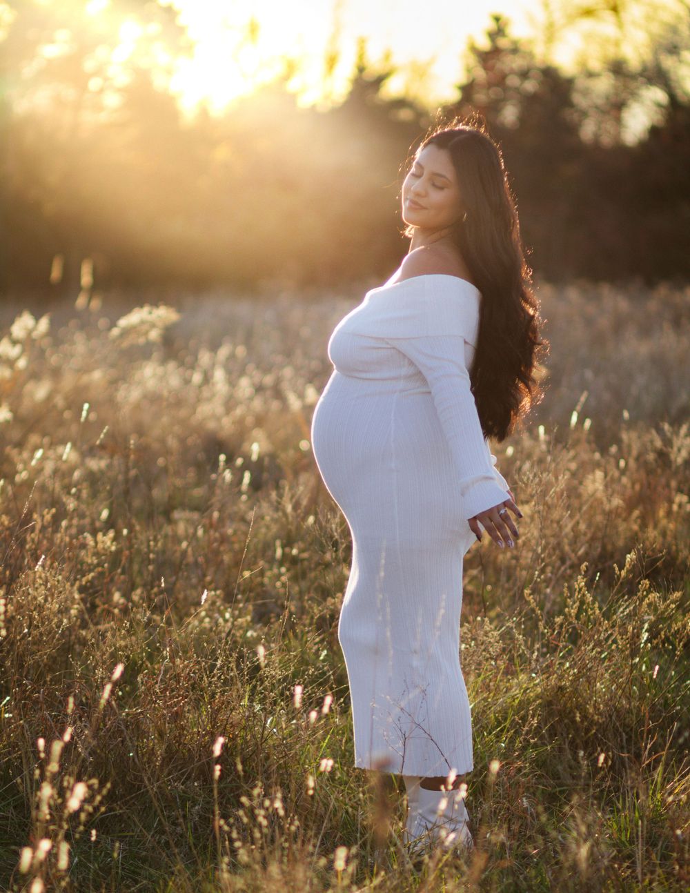 A pregnant woman in a white dress stands in golden sunlight amid tall grass during sunset.