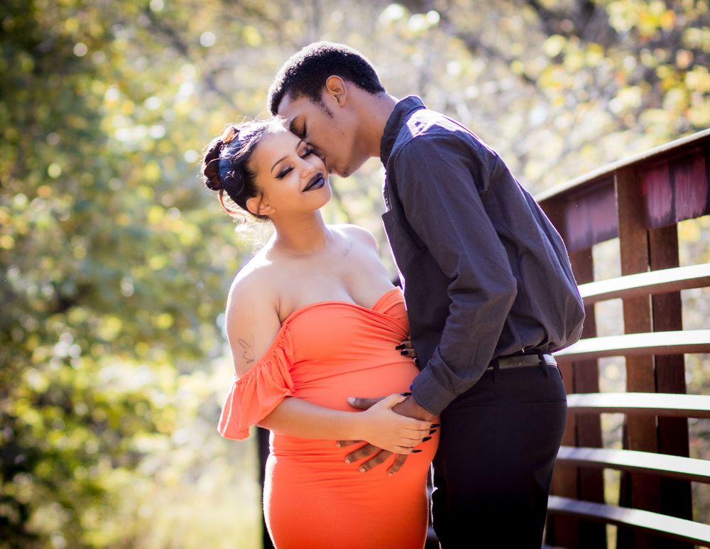 A romantic couple embraces on a wooden bridge, with the woman wearing a bright coral dress against a blurred nature background.