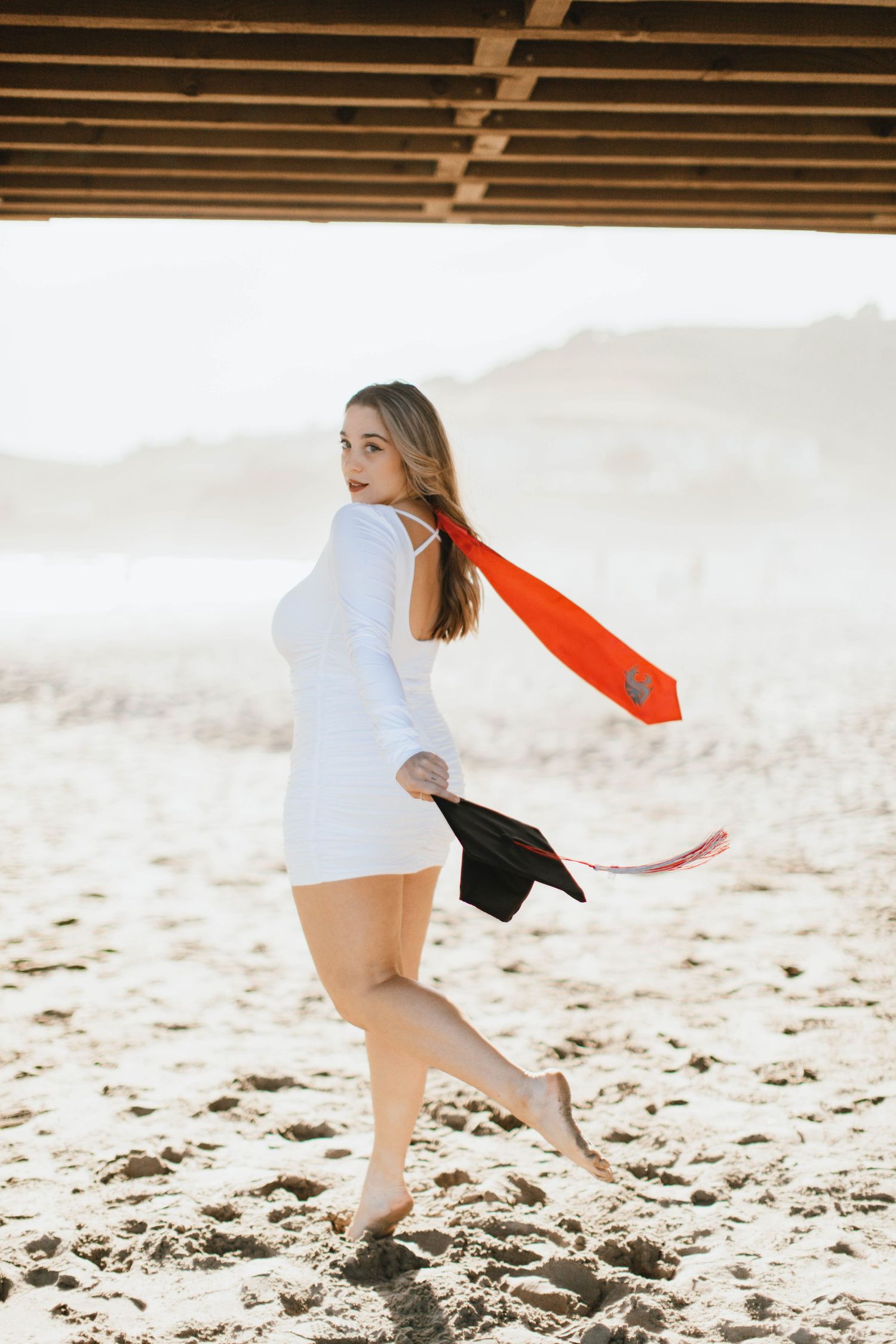 Dynamic Graduation beach photoshoot under a pier wearing white and red flowing fabrics.