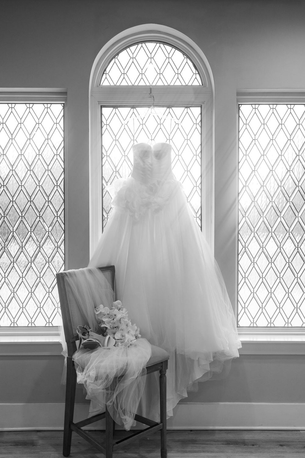 An elegant white wedding dress hangs by a window with diamond-patterned glass and a chair adorned with flowers.