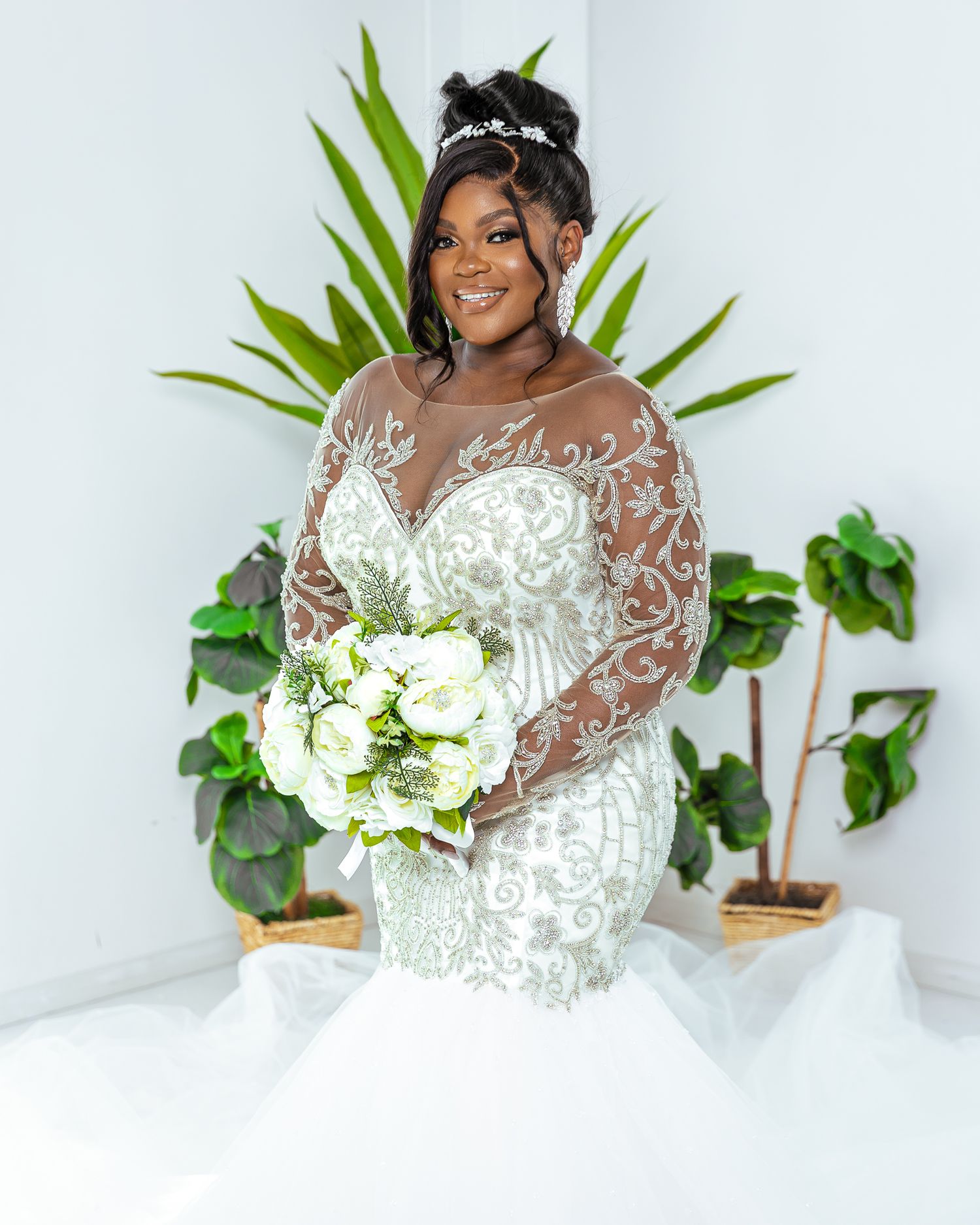 Bride in lace wedding gown holding white bouquet poses in front of tropical green plants.