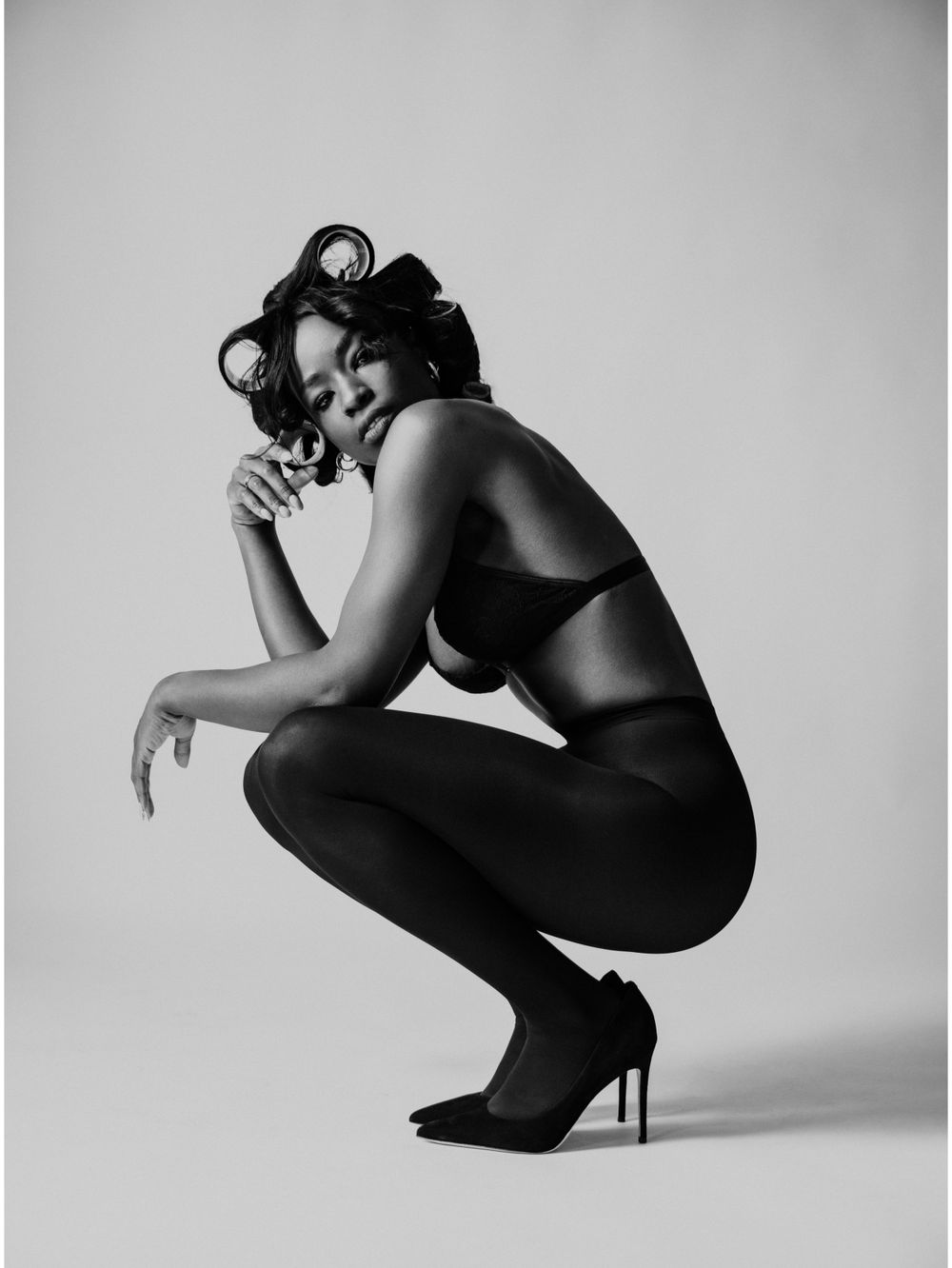 Artistic black and white portrait of a figure in lingerie striking a crouching pose with vintage-style hair.