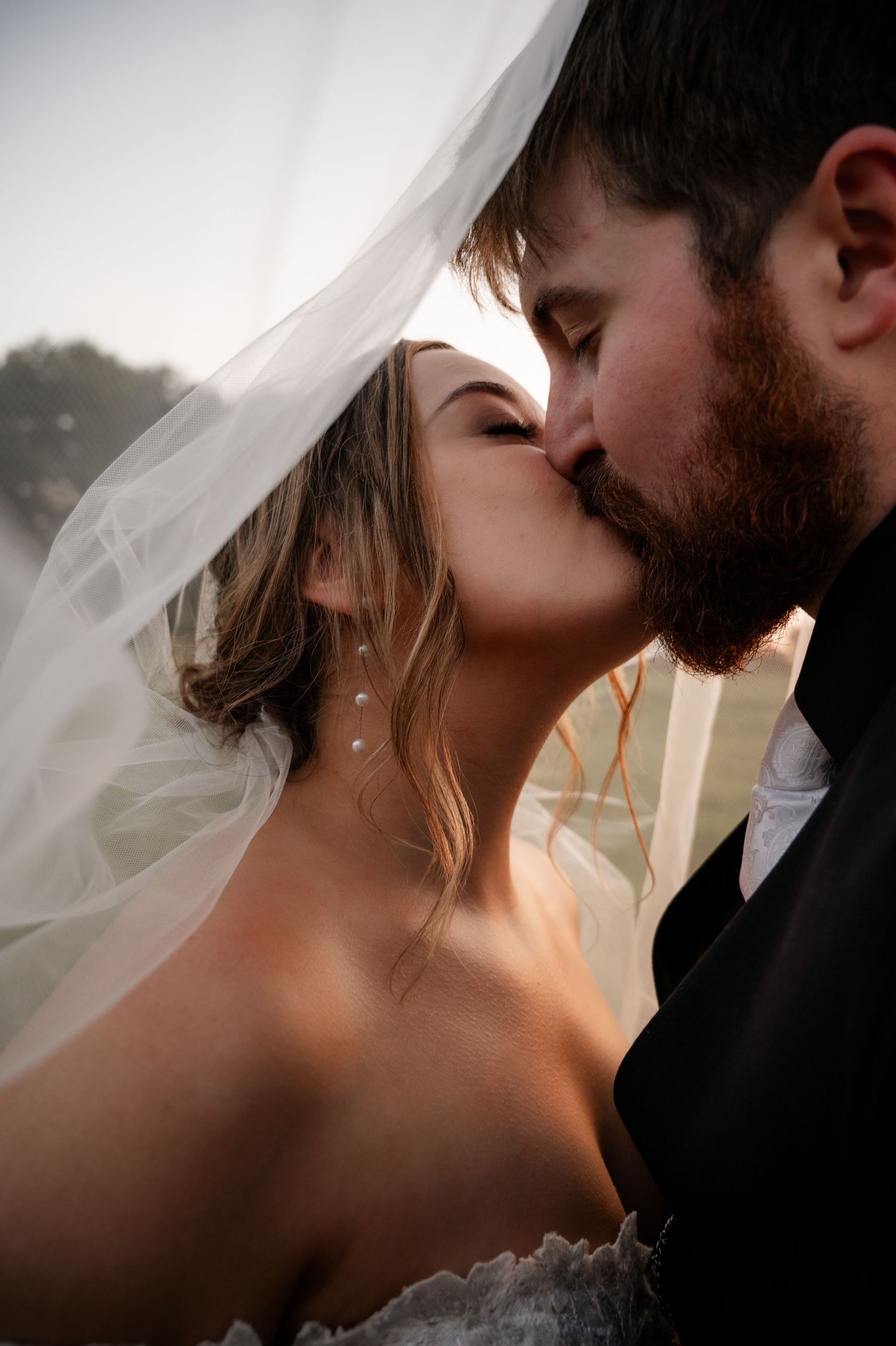 Bride and groom sharing romantic kiss under flowing veil in warm golden wedding portrait.