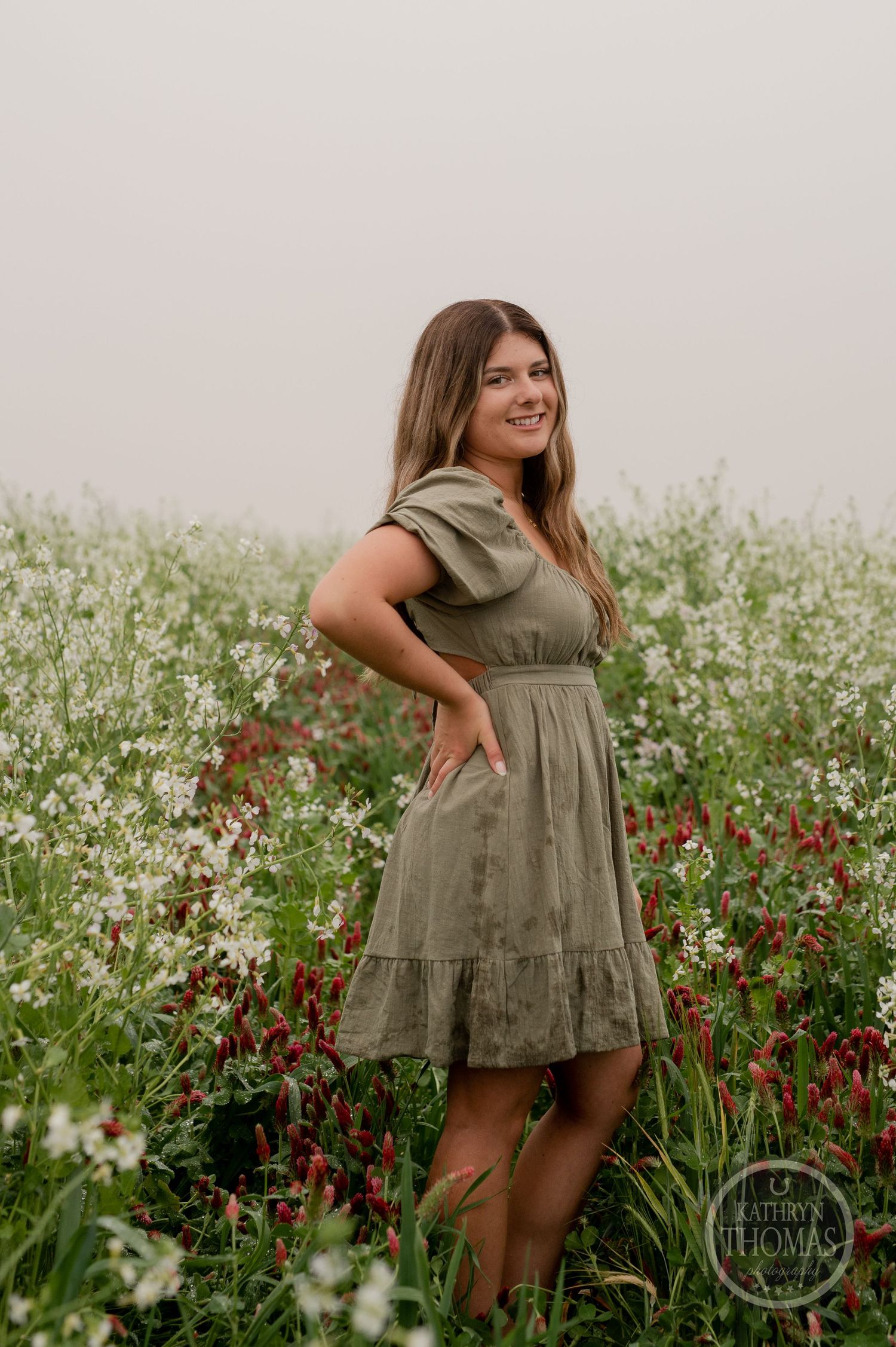 Person in olive dress smiles joyfully while posing in a meadow with red and white flowers.