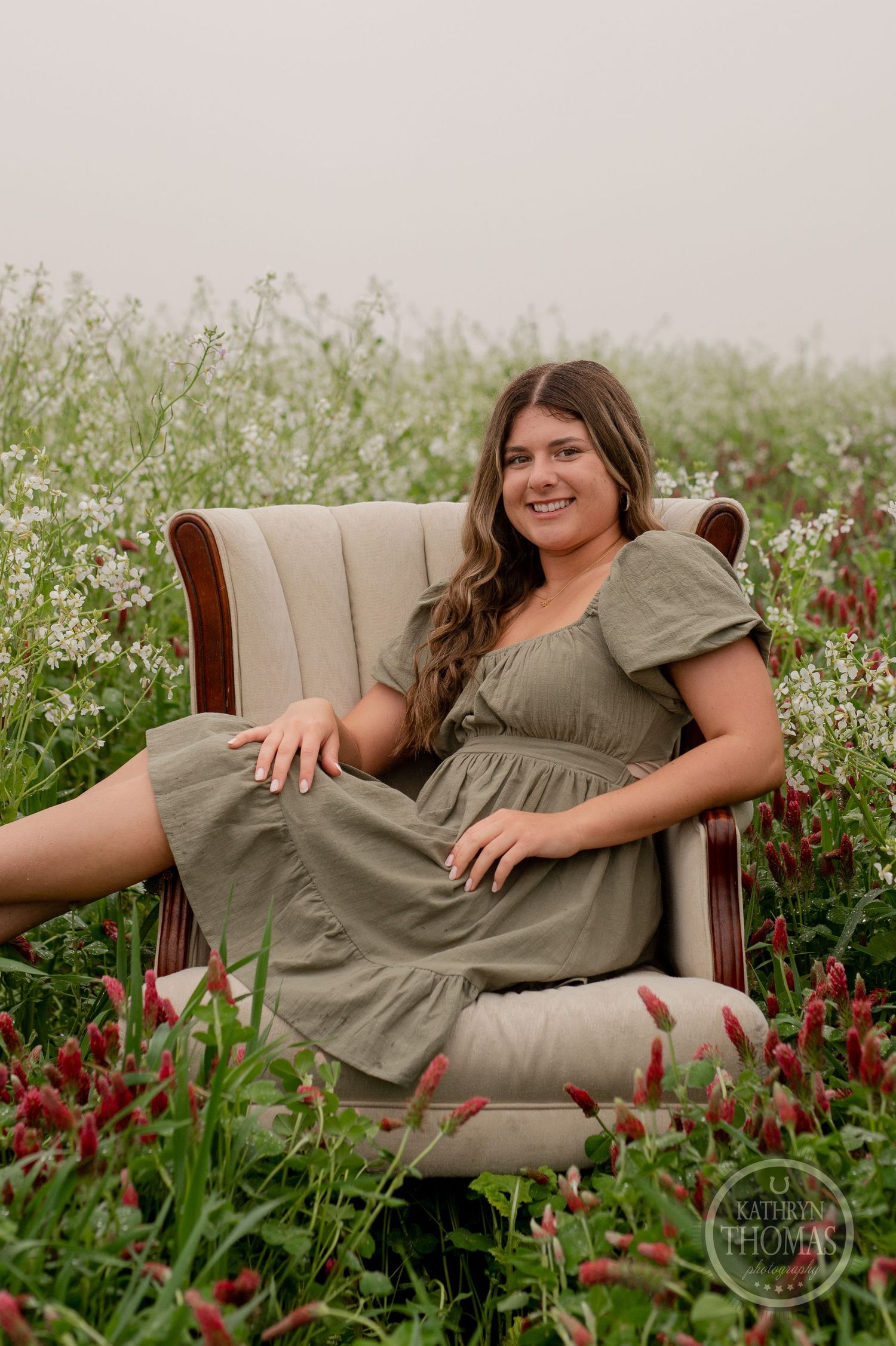 A woman in an olive dress sits comfortably in a vintage armchair among blooming wildflowers.