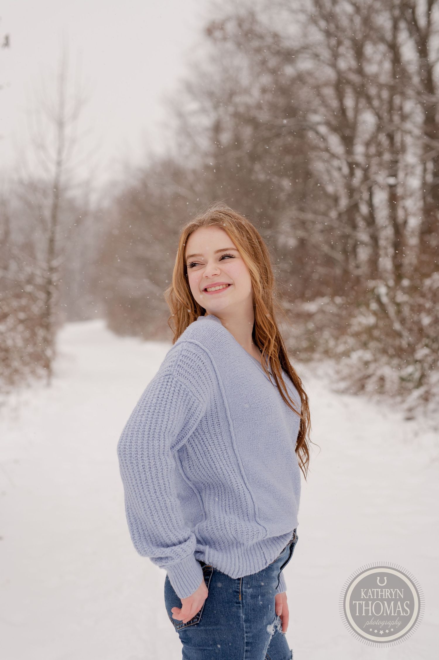 A casual winter portrait featuring someone in a blue sweater and jeans against a snowy backdrop.