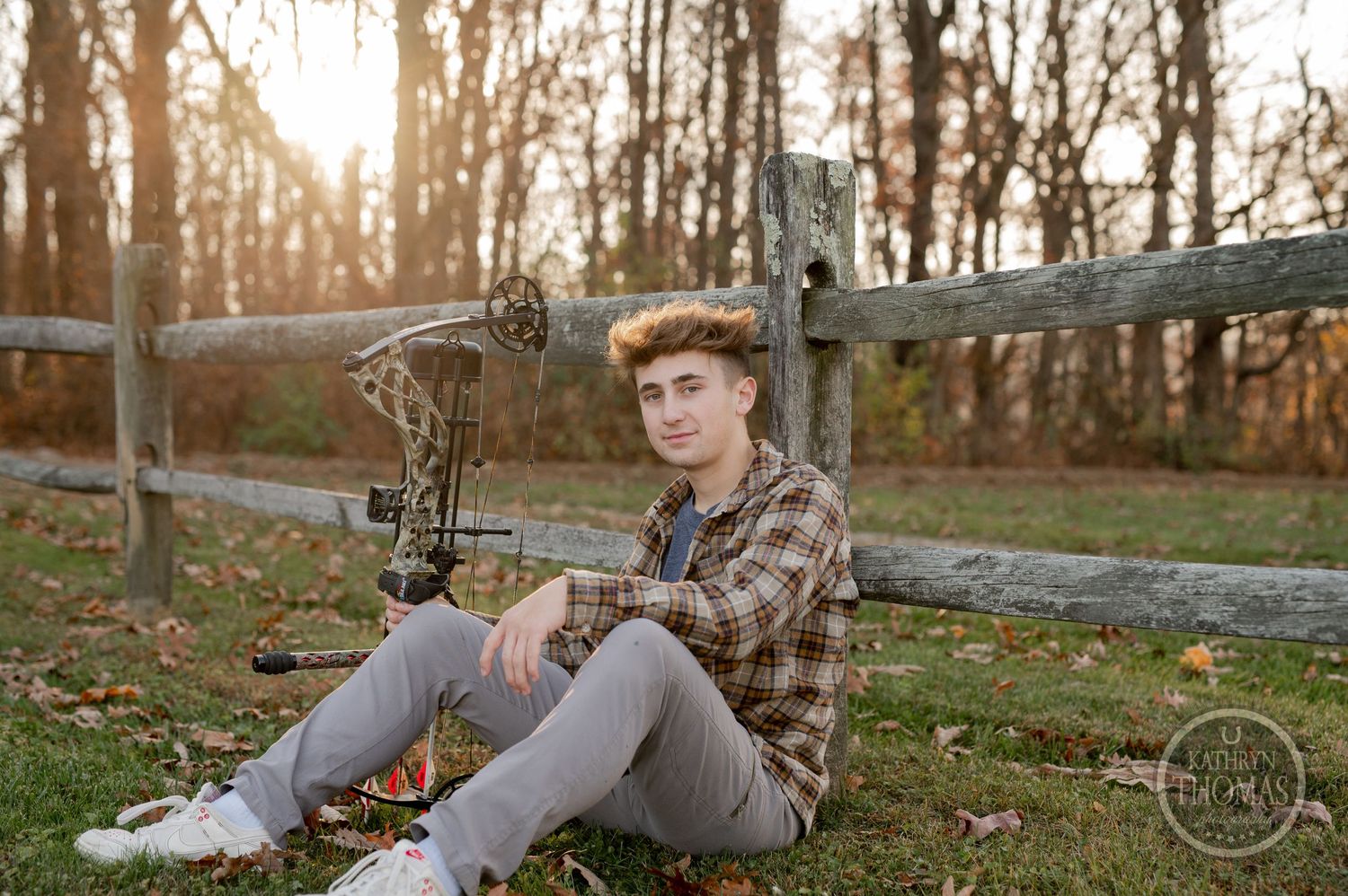 Someone in plaid sits by a wooden fence with a compound bow during golden hour.