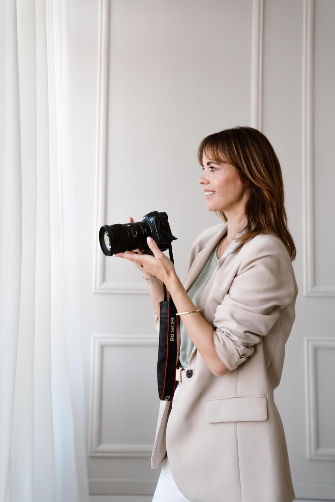 Professional photographer in light beige blazer holding camera and smiling against white wall with molding.