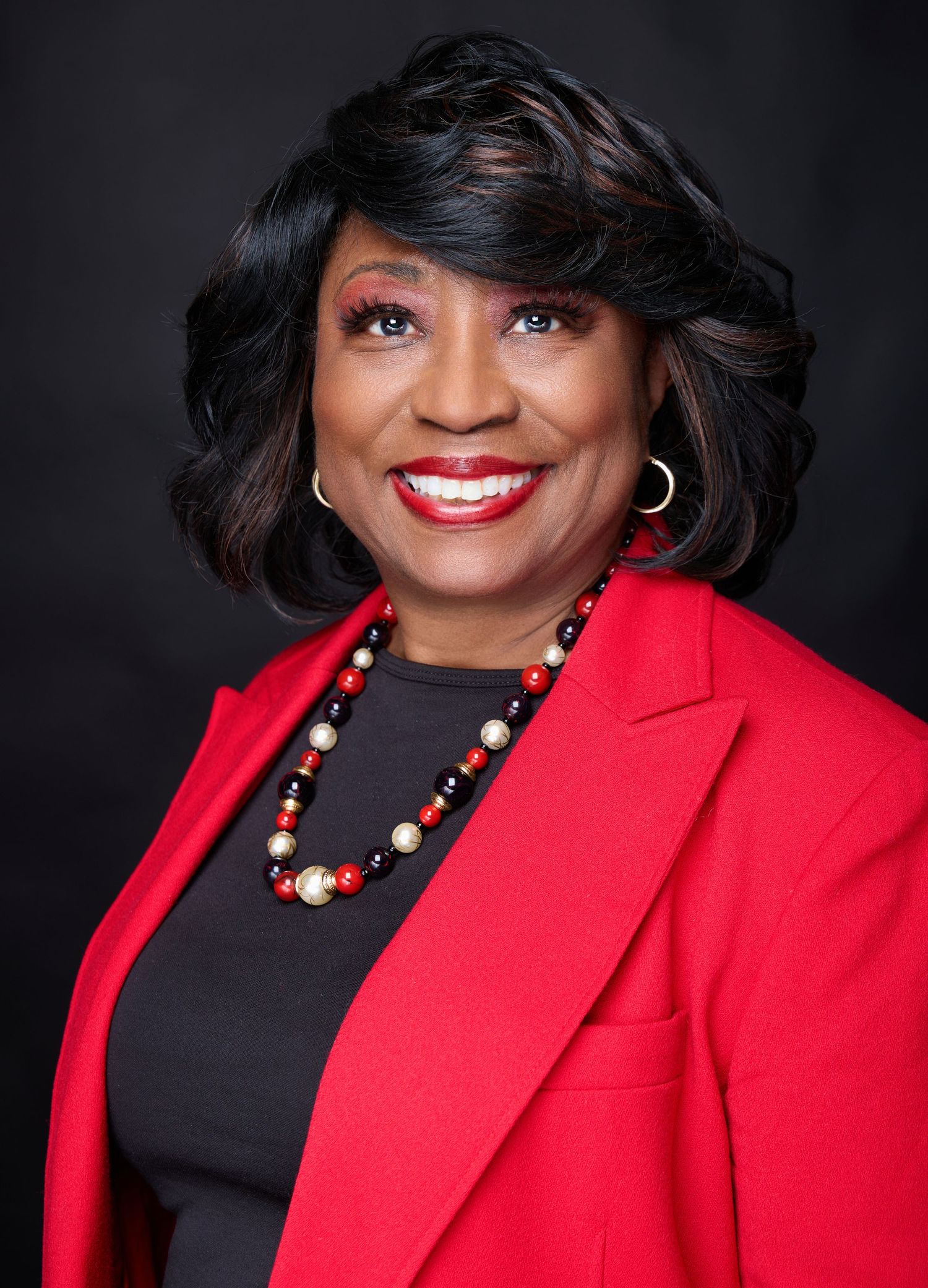 Professional headshot portrait in red blazer and black top with statement necklace against dark background.