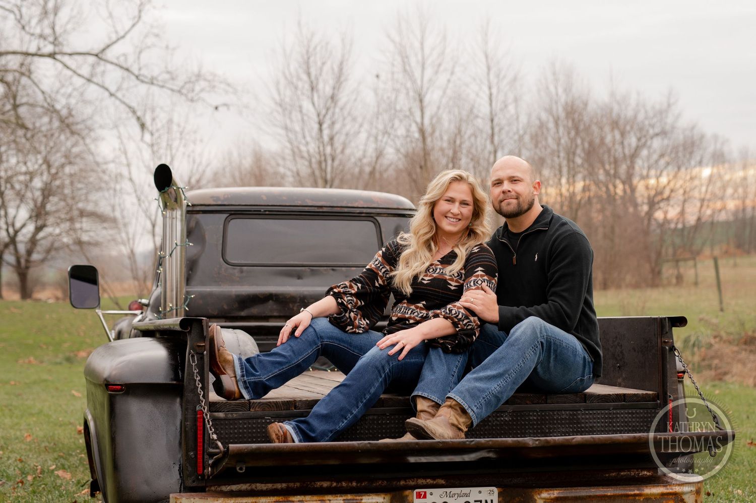 Couple cuddling with dog in back of vintage pickup truck in rural setting.
