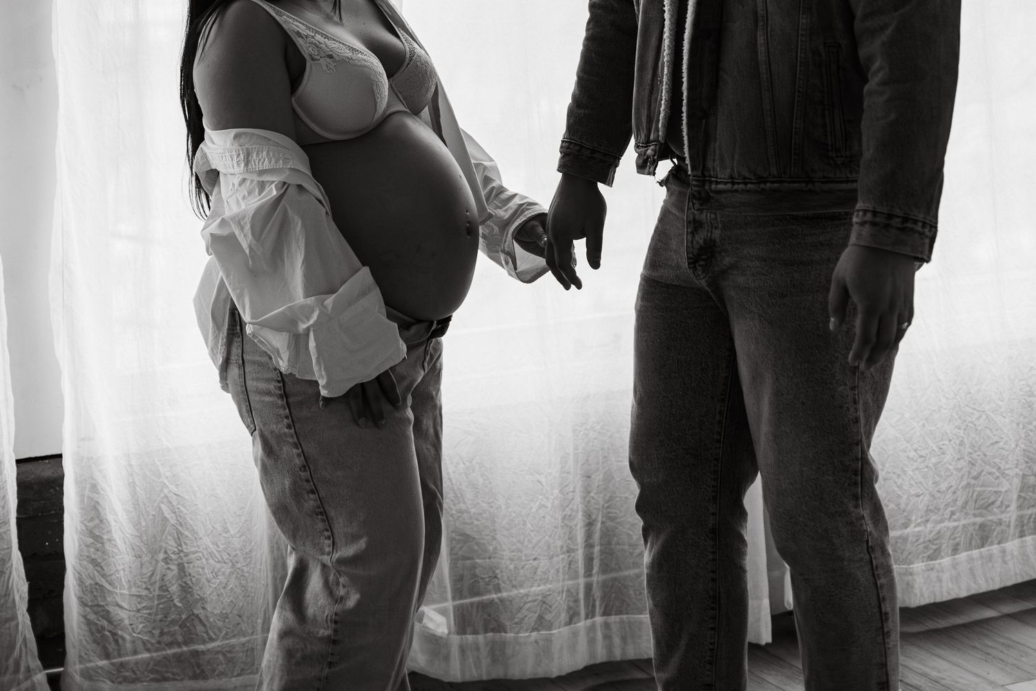 A black and white photograph showing two people standing and holding hands against a plain light background.