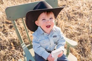 Small child in cowboy hat and denim shirt sitting on green chair outdoors.
