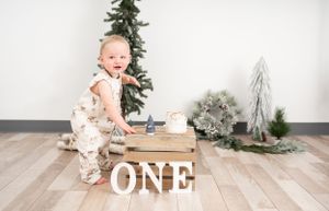 Child stands next to wooden ONE letters with Christmas decorations and trees in background.