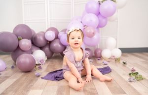 Baby in pink outfit sits on floor surrounded by purple and pink balloons for first birthday celebration.