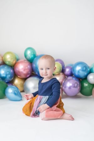 Baby in navy and orange dress sitting with colorful balloon arch background.