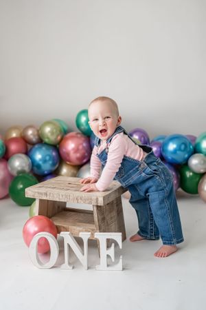 First birthday celebration setup with colorful balloons and wooden stool against white backdrop.