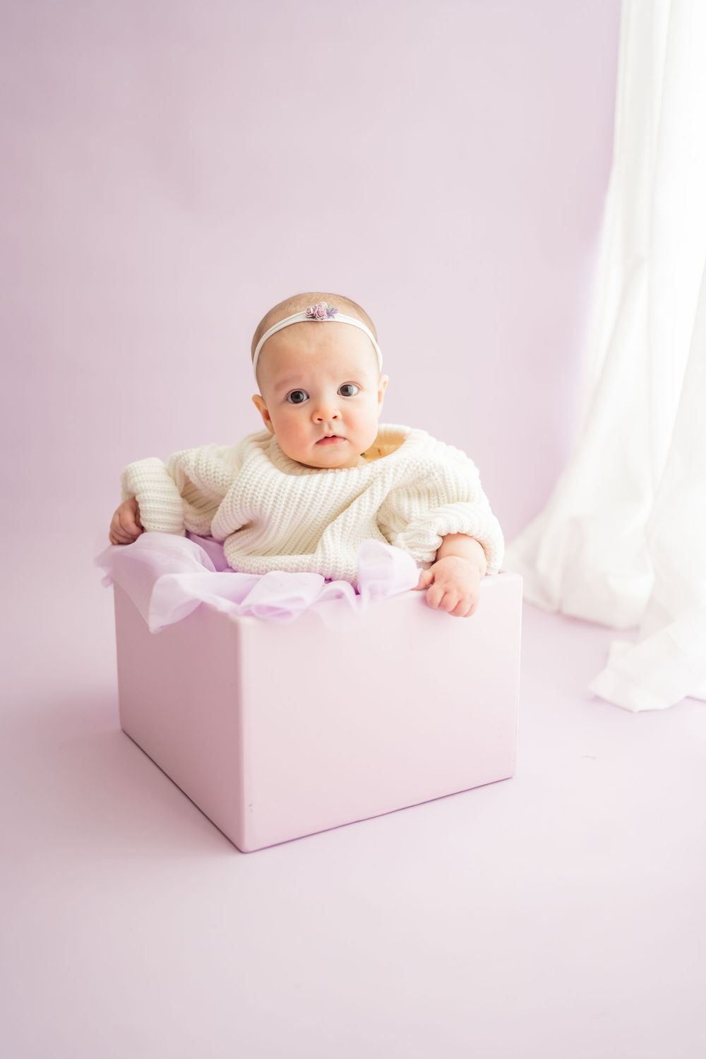 Baby in cream sweater resting in pink box during studio photography session.