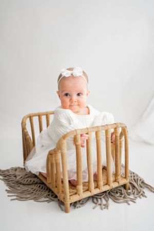 Baby in white outfit sitting in rustic bamboo crib on textured grey blanket.