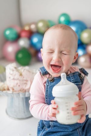 Baby in denim overalls enjoying a bottle with colorful balloons and cake in background.