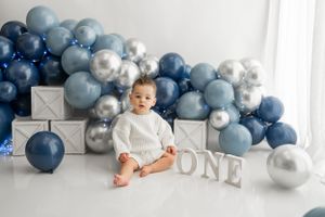 First birthday setup with blue and silver balloons and white 'ONE' letters against white backdrop.