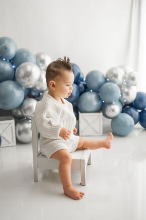 Small child in white sweater sitting on chair with balloon decoration background.