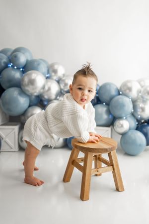 Little one in white sweater leaning on wooden stool with blue balloon backdrop.