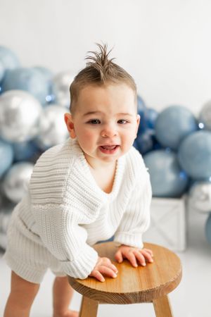 Toddler in white sweater smiling while leaning on wooden stool.