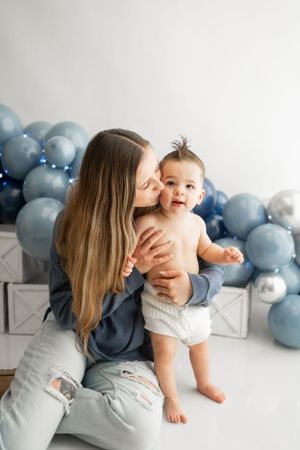 Sweet moment with mother and baby against blue balloon backdrop.