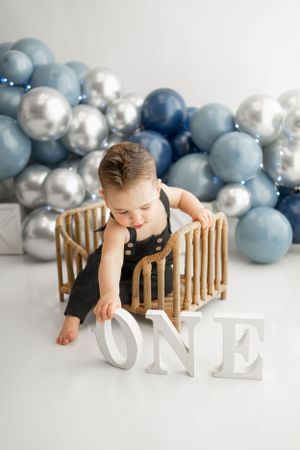 Baby in dark overalls reaching for white 'ONE' letters with balloon background.