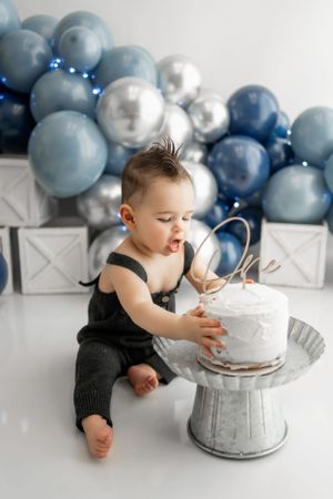 Baby exploring white cake on metal pedestal with blue and silver balloon backdrop during photoshoot.