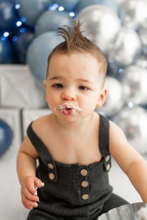 Cake smash portrait with messy face against blue and silver balloon backdrop.