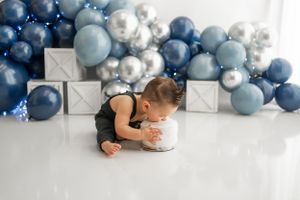 Small child in black outfit explores white cake against blue and silver balloon backdrop during cake smash photoshoot.