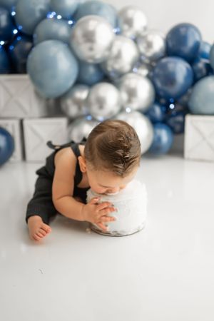 Baby laying on floor investigating small cake with decorative blue and silver balloons in background.