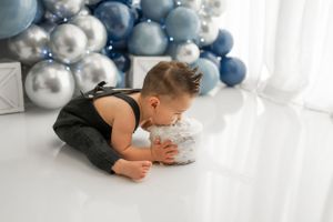 Baby in black outfit eating cake during first birthday photoshoot with blue balloon arch.