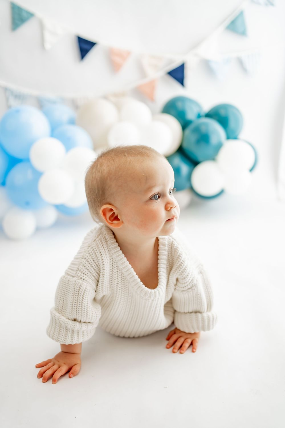 Small baby in white knit sweater crawling on floor with blue and white balloon decorations and bunting in background.