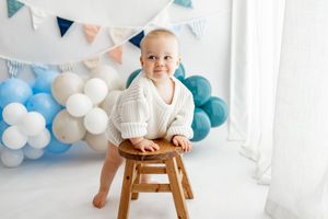 Baby wearing white clothes sits on wooden stool with blue and white balloon garland decorations in background.