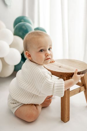 Baby in white knit sweater sits beside wooden stool with green and white balloons in background.