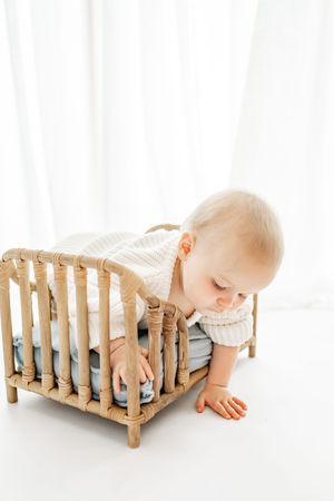 Baby in white sweater leaning over wooden crib railing in bright minimalist setting.